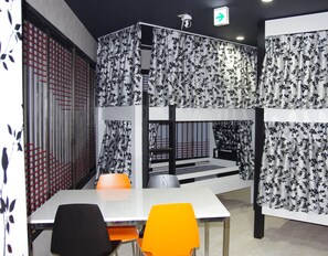 A bunk bed room with a white table and orange chairs, a security camera, and a directional sign. - Sora Hostel (Osaka)