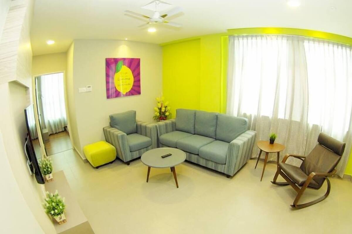 Un salon avec un canapé gris, une petite table ronde, un fauteuil à bascule en bois et un pouf jaune. Il y a un tableau au mur et un ventilateur de plafond.