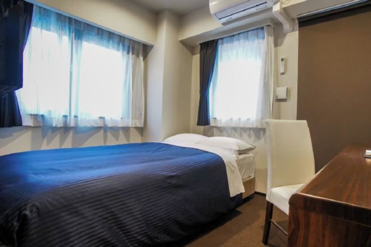 Hotellihuone, jossa on sänky, tuoli, pöytä ja kaksi ikkunaa, joissa on verhot.
