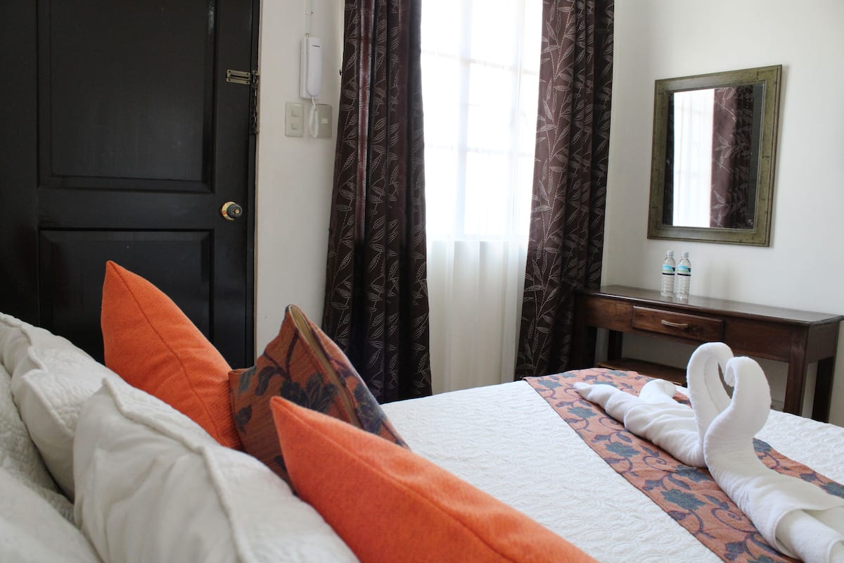 Habitación de hotel con cama, almohadas, escritorio, espejo y ventana con cortinas.