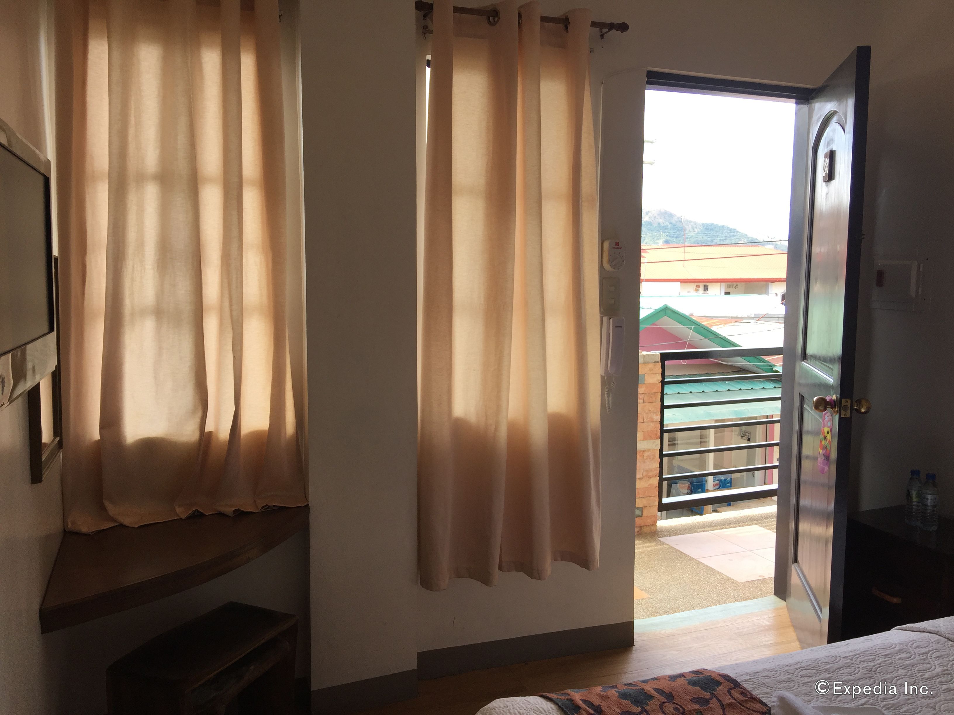 A hotel room with a bed, a desk, a chair, and a window with curtains. There is a door leading outside.