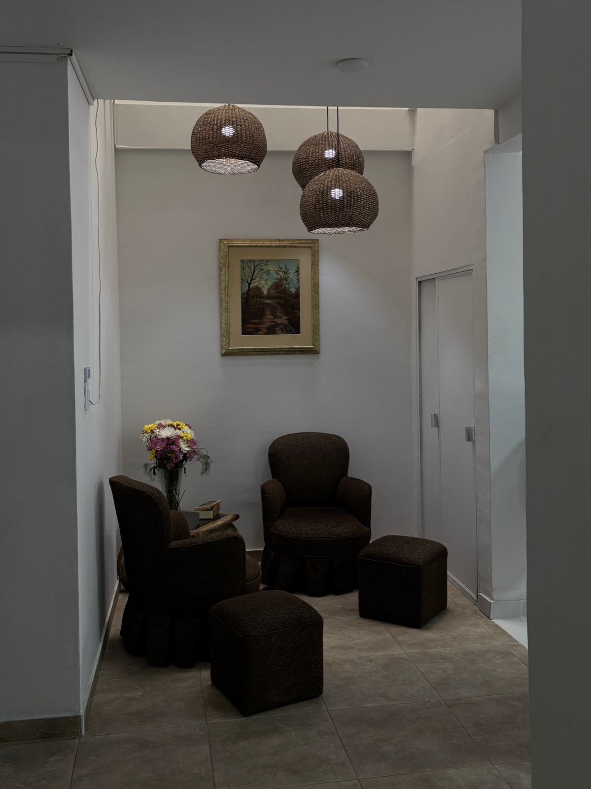 A hallway with a dark brown armchair, a small table with a vase of flowers, and three hanging pendant lights.