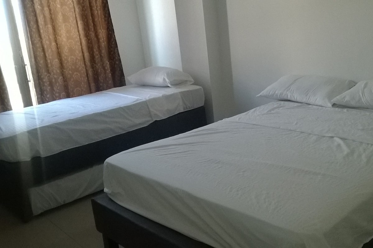 Two single beds with white linens in a room with a plain white wall and a window with curtains.