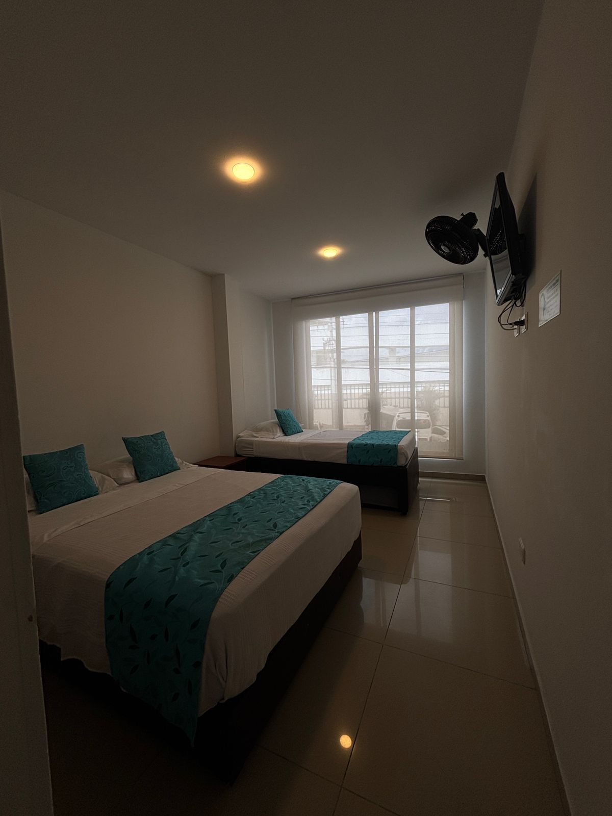 A hotel room with two beds, a large window, a ceiling fan, and a flat-screen TV mounted on the wall.