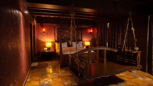 Um quarto com pouca luz, com uma cama grande, uma cadeira de madeira e uma pequena mesa.