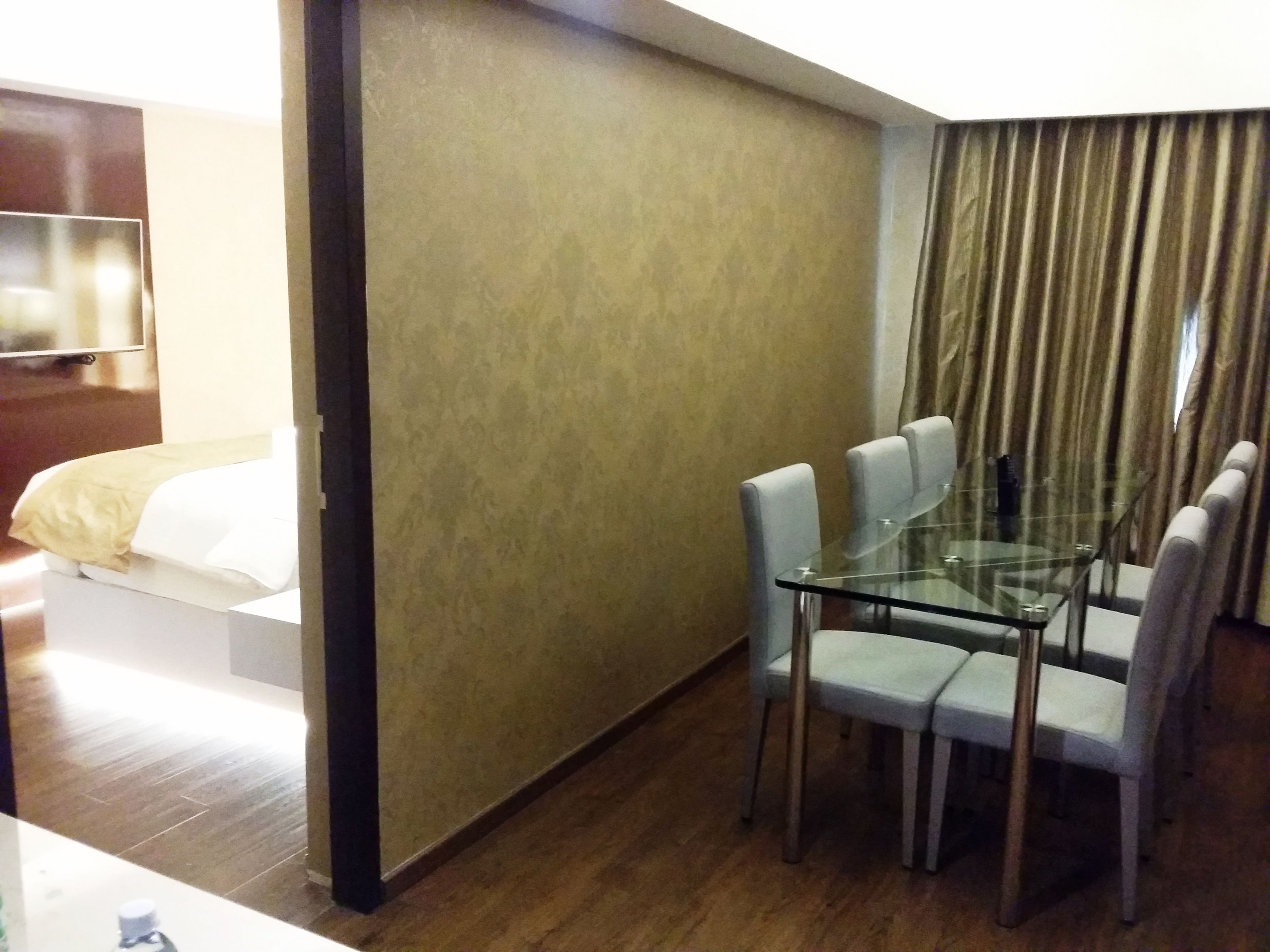 A hotel room with a glass dining table, four chairs, a bed with white bedding, a television, and a curtain.