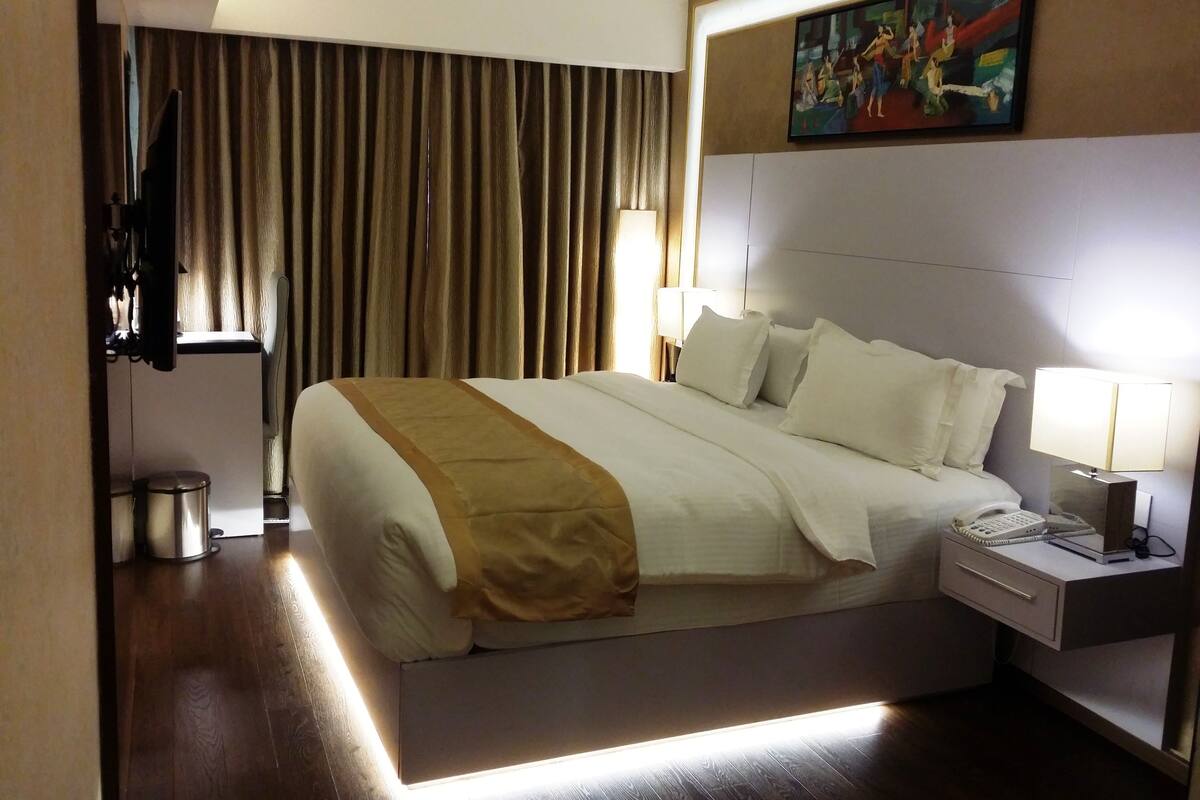 Habitación de hotel con una cama grande, una mesita de noche con lámpara, un cuadro en la pared y una cortina.