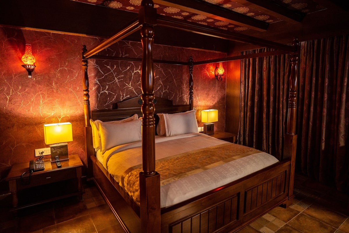 Una cama con dosel de madera, dos mesitas de noche con lámparas, y un papel tapiz estampado.