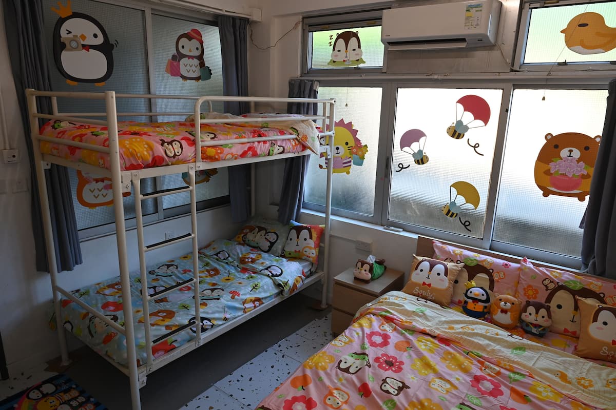 A bunk bed room with a window view, a bedside table with a teddy bear, and a bed with a floral bedspread.