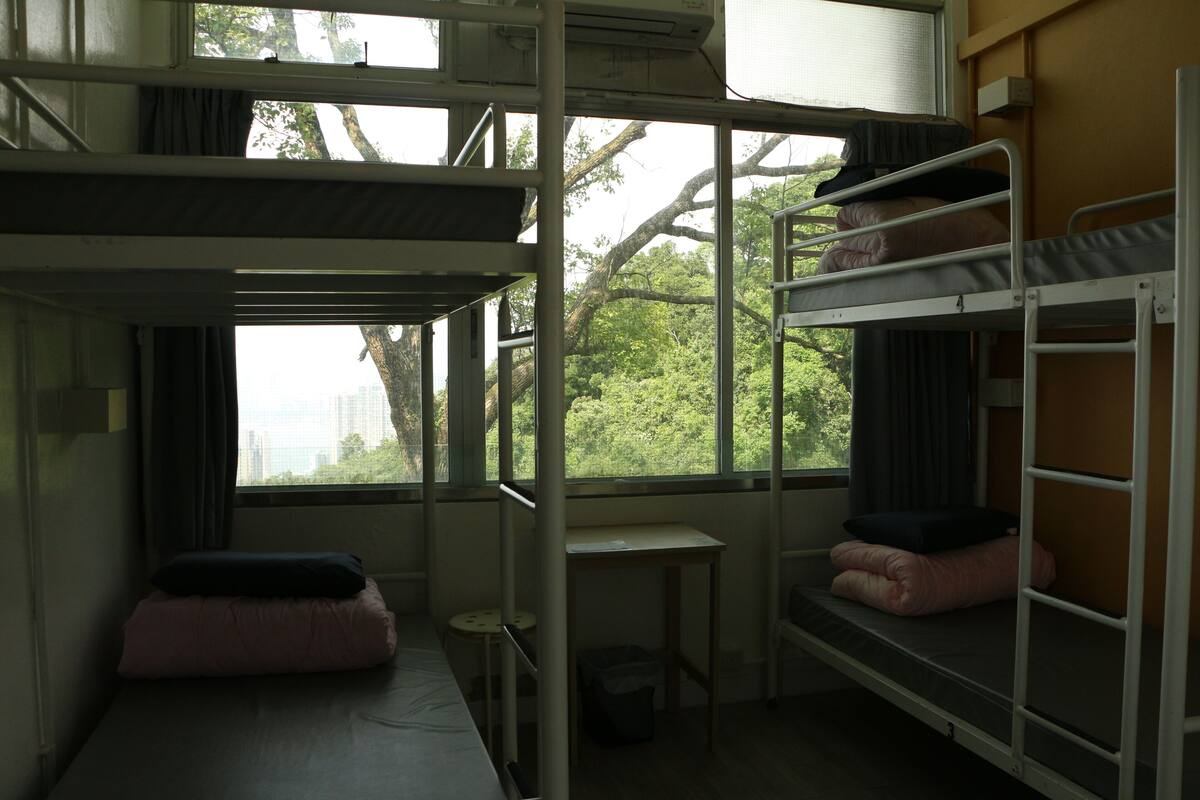 A dormitory room with bunk beds, a small table, and a window with a view of trees and buildings.