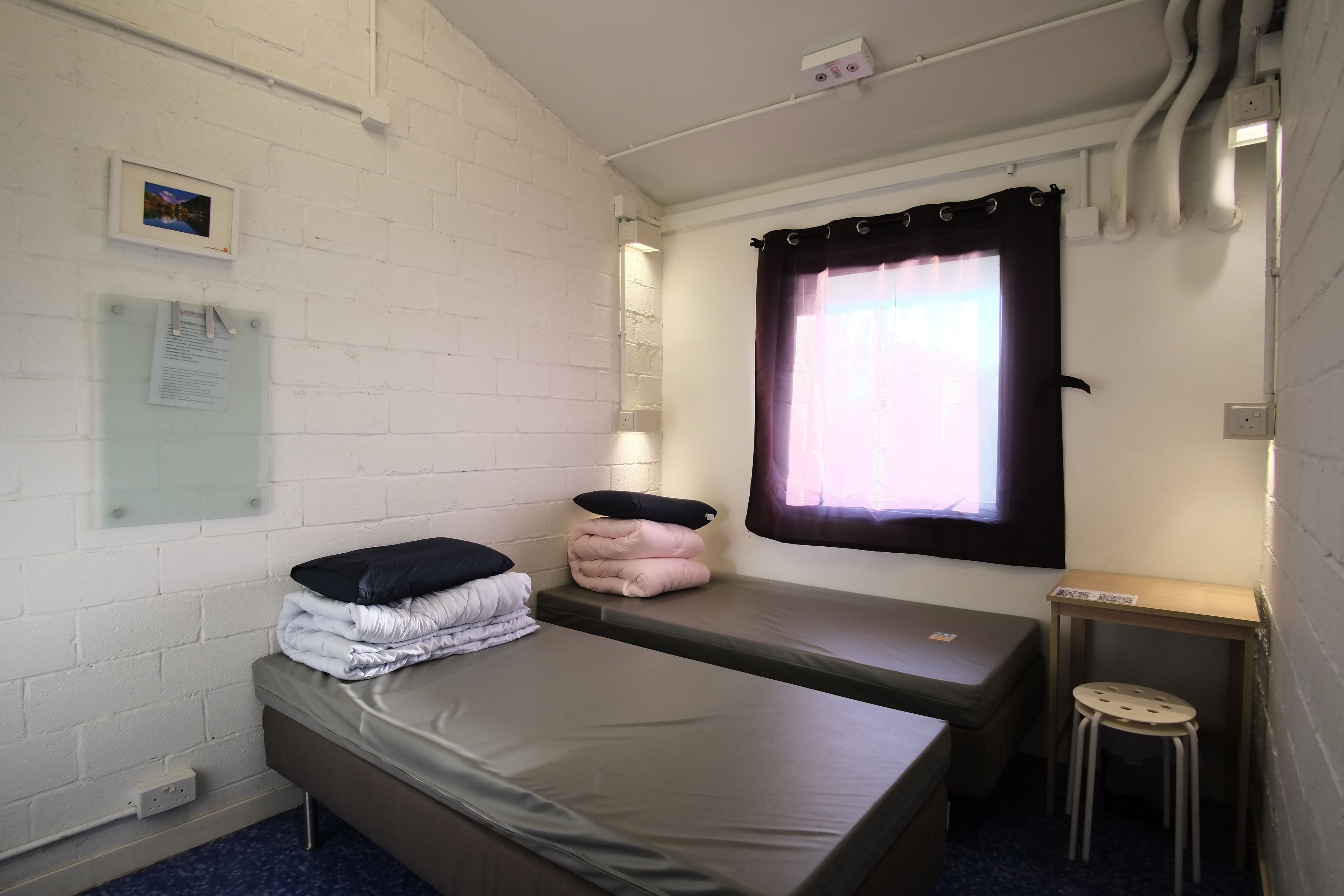 A small, compact room with two single beds, a small table, and a window with curtains.