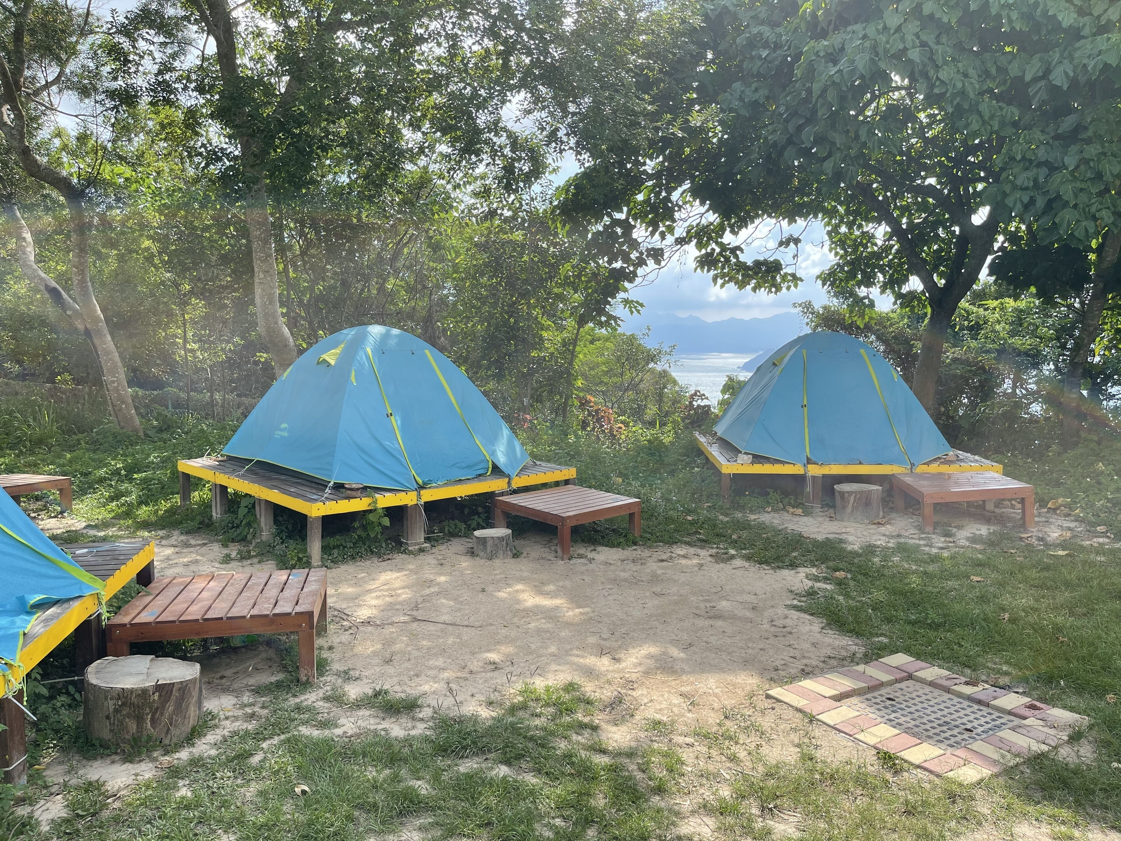 Zwei blaue Zelte mit gelben Fundamenten, Holzbanken und ein Holztisch auf einem Campingplatz mit Bäumen und Blick auf das Wasser.