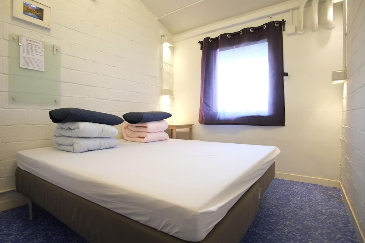 A small, neatly made bed with white bedding and two pillows, a small wooden nightstand, a window with a dark curtain, and a white brick wall with a framed picture and a notice board.