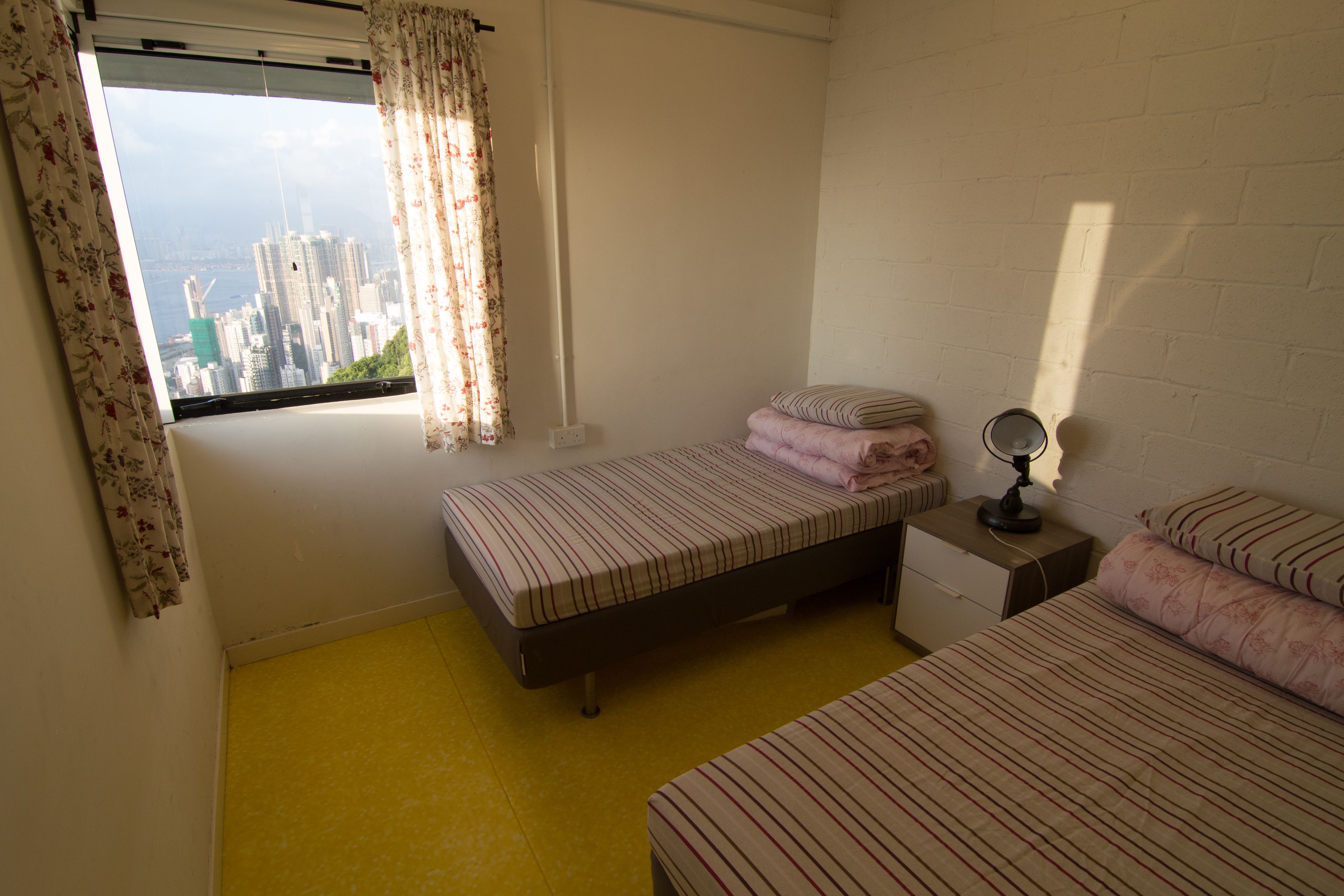 A bedroom with two beds, a window with curtains, a bedside table with a lamp, and a view of a cityscape.