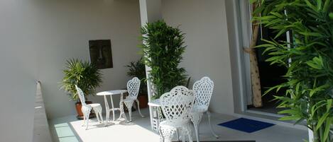 A covered outdoor seating area with white metal chairs and tables, surrounded by potted plants.
