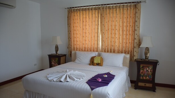 A neatly made bed with a white comforter and pillows, a decorative pillow, a purple towel, and a small elephant plush toy. There are two bedside tables with lamps and a decorative item on one. - Sumali Villa (Sing Buri)