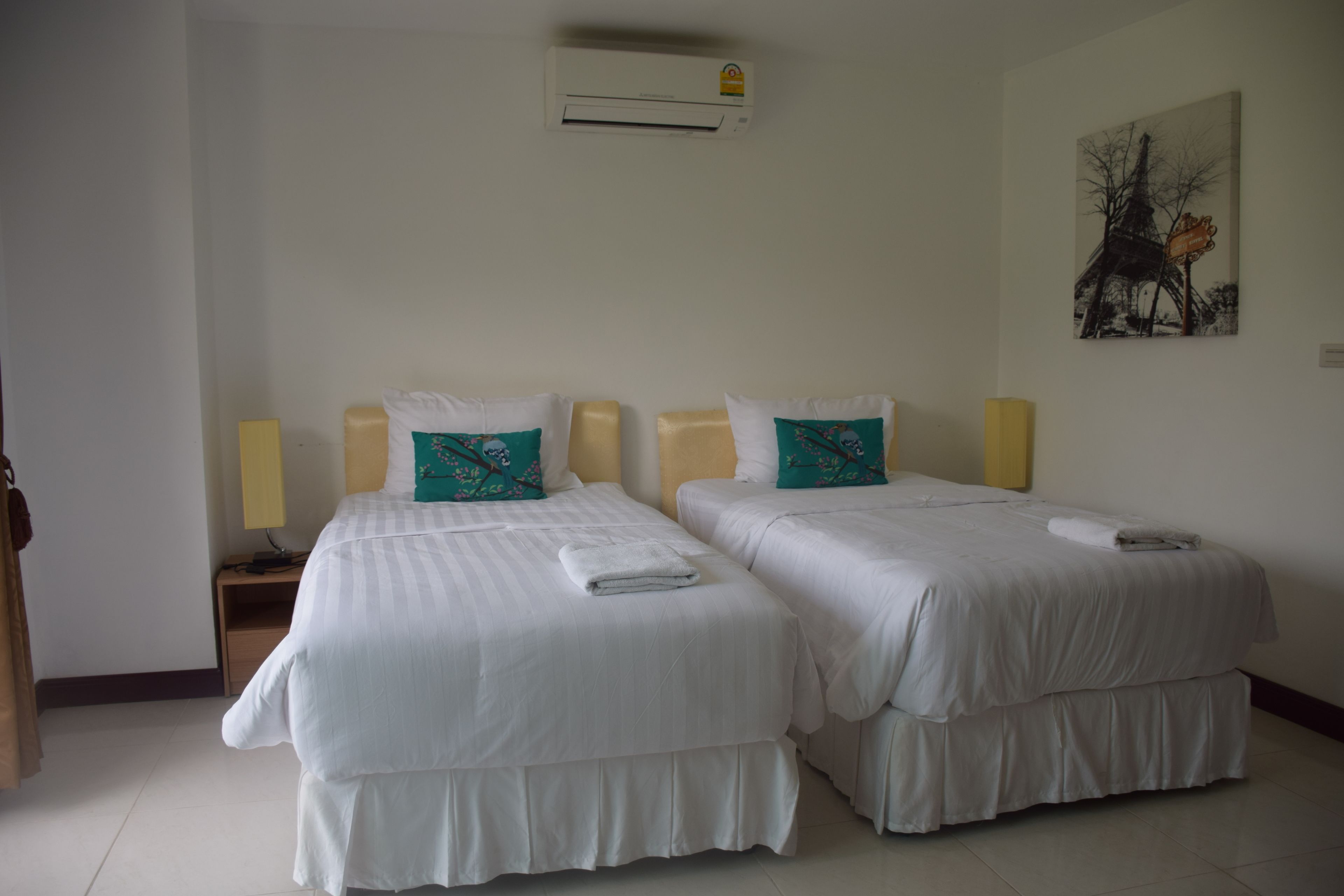 A hotel room with two beds, each with white bedding and teal pillows, a nightstand with a lamp, and a painting on the wall.