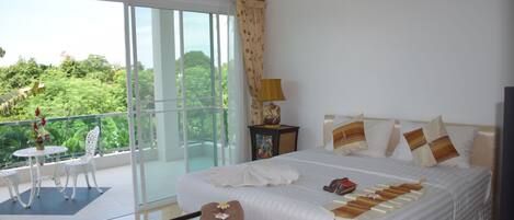 Quarto de hotel com uma cama grande, varanda com mesa e cadeiras, e vista para o verde lá fora.