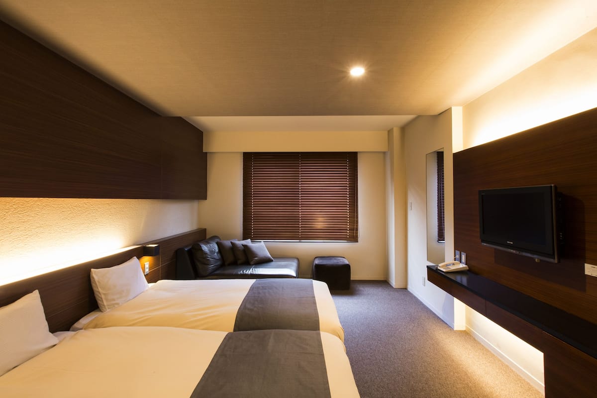 Une chambre d’hôtel avec deux lits, un téléviseur à écran plat, une fenêtre avec des stores et un petit coin salon.