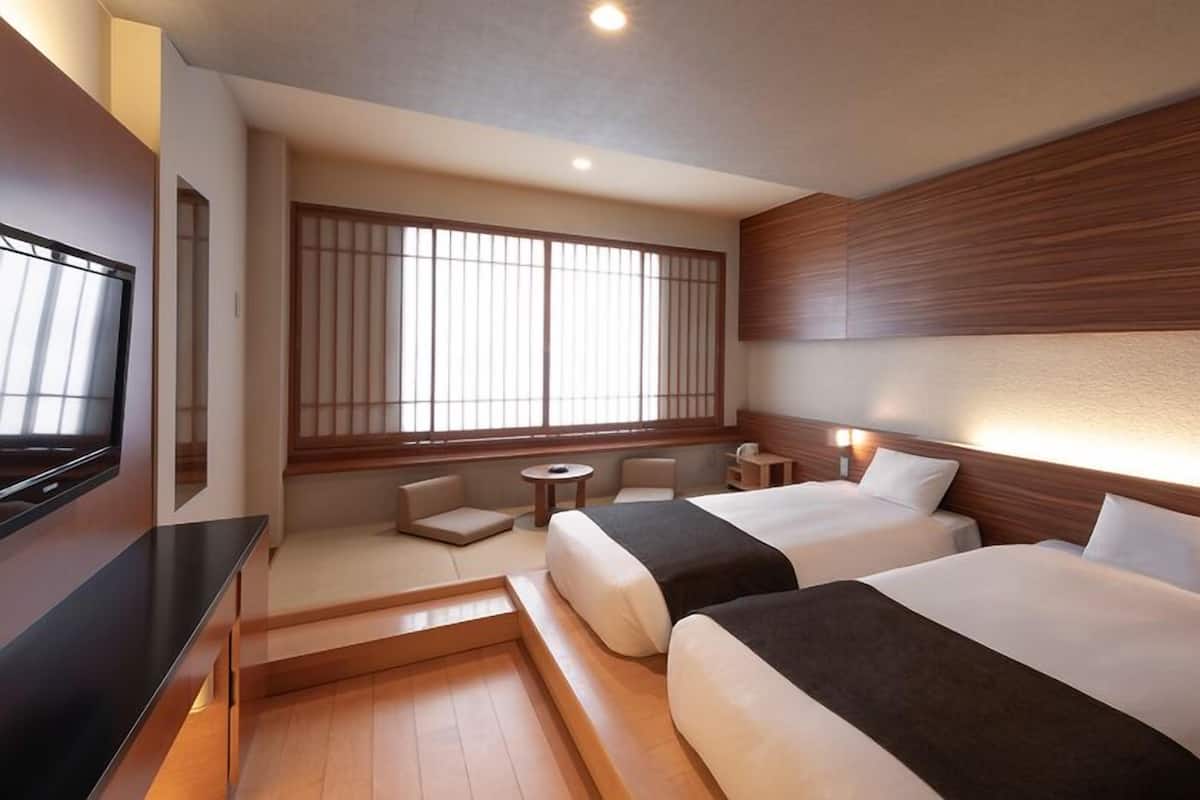 Une chambre d’hôtel avec deux lits, une télévision, un coin salon et une grande fenêtre avec des stores.