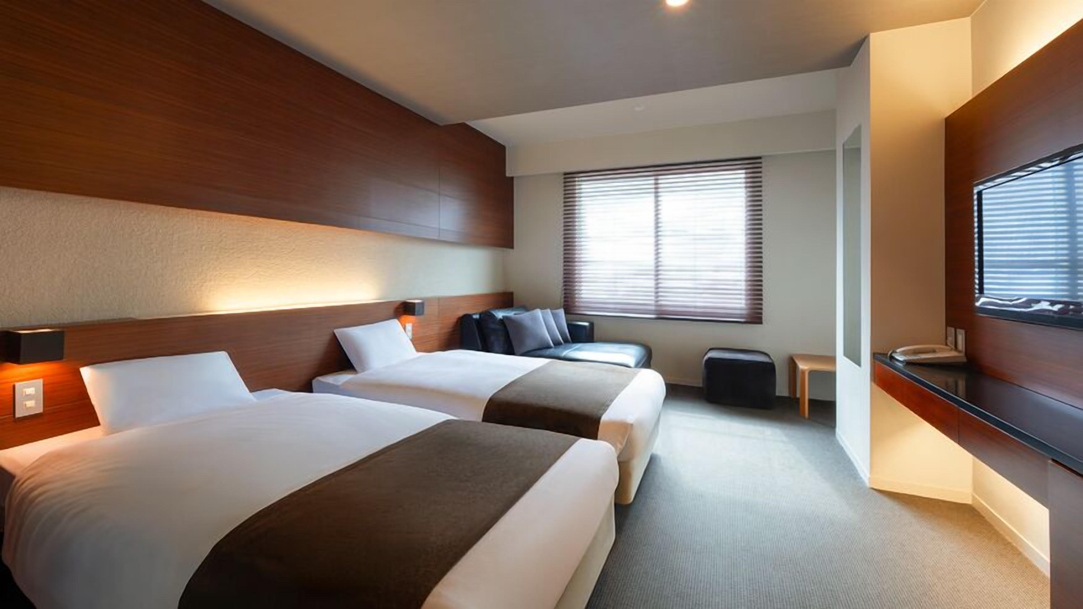 Une chambre d’hôtel avec deux lits, un petit coin salon, une télévision et une fenêtre avec des stores.