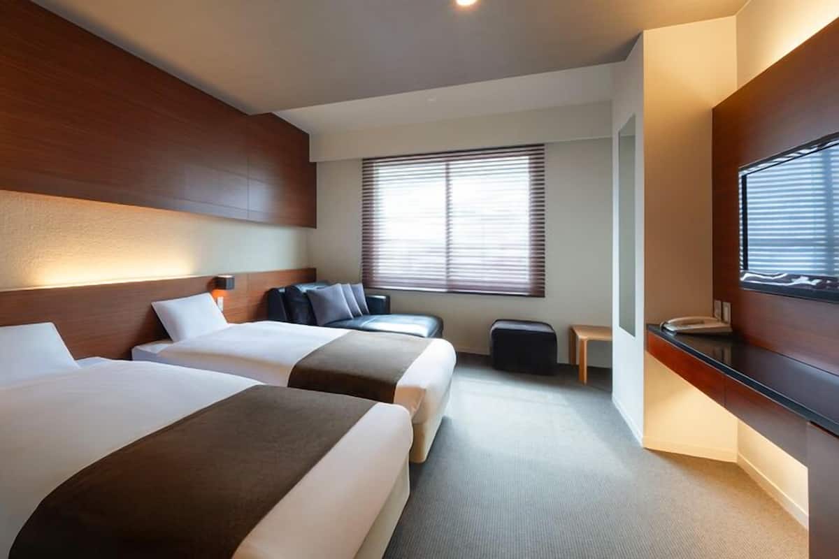 Une chambre d’hôtel avec deux lits, un petit coin salon, une télévision et une fenêtre avec des stores.