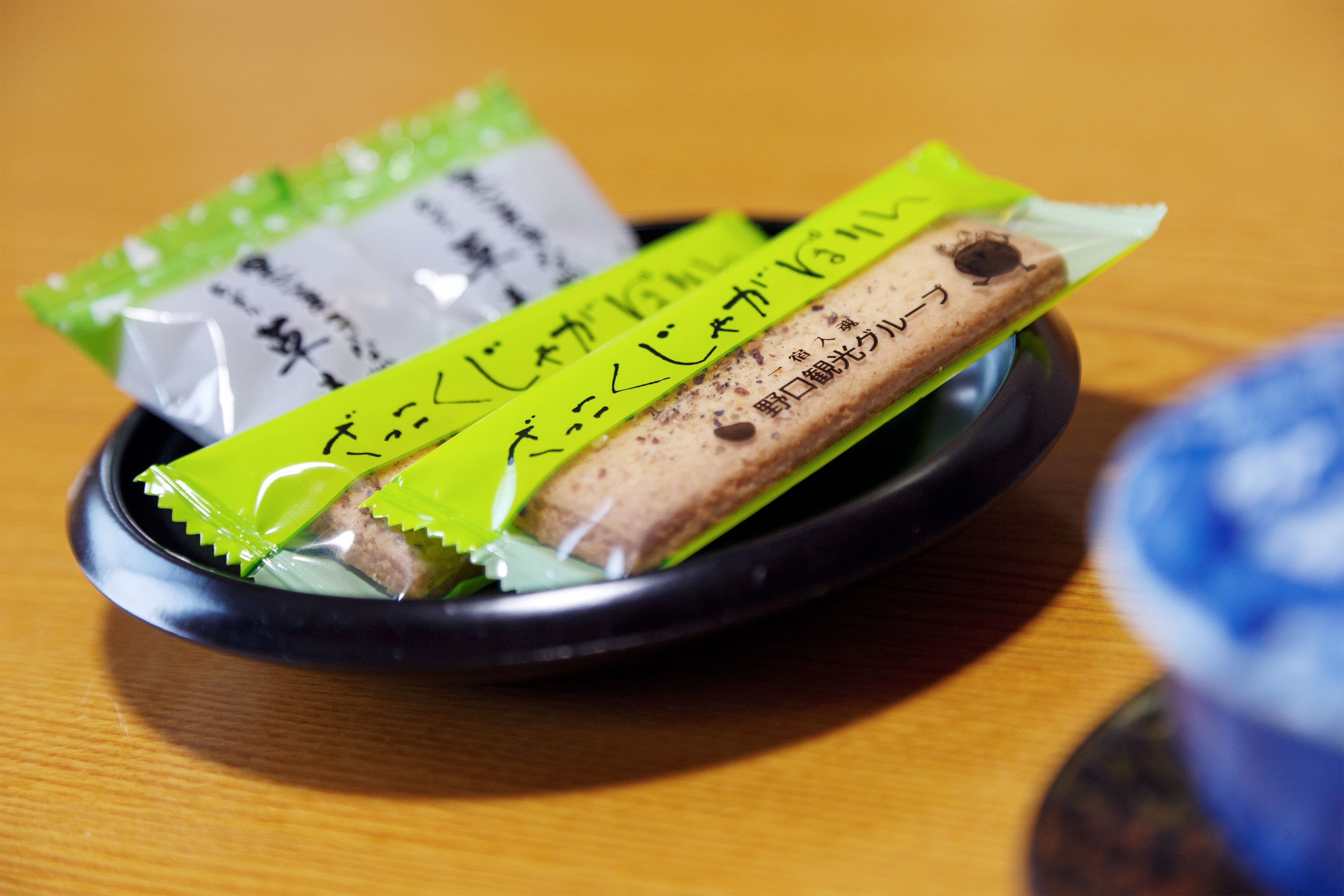 A black bowl containing a wrapped food item and a green packet with Japanese text, placed on a wooden surface.