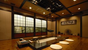 Une pièce de style japonais traditionnel, avec un sol recouvert de tatamis, une petite table agrémentée d’un élément décoratif aquatique et une grande fenêtre donnant sur un paysage verdoyant.
