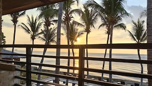 Un balcón con una mesa de vidrio y una taza negra, con vista a una puesta de sol, palmeras y el océano.