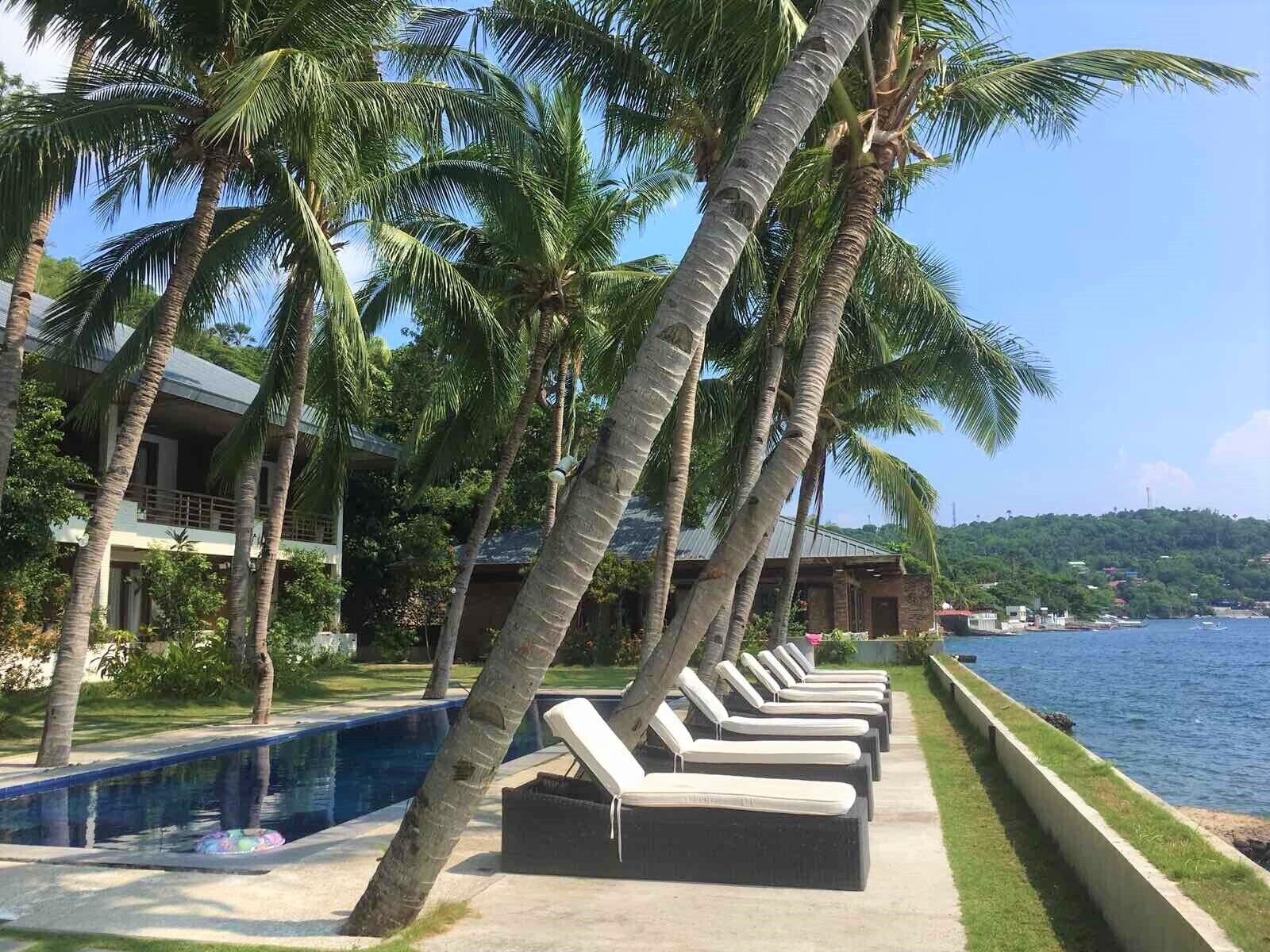 Zona de piscina con sillones de descanso y palmeras, con vista a una masa de agua.