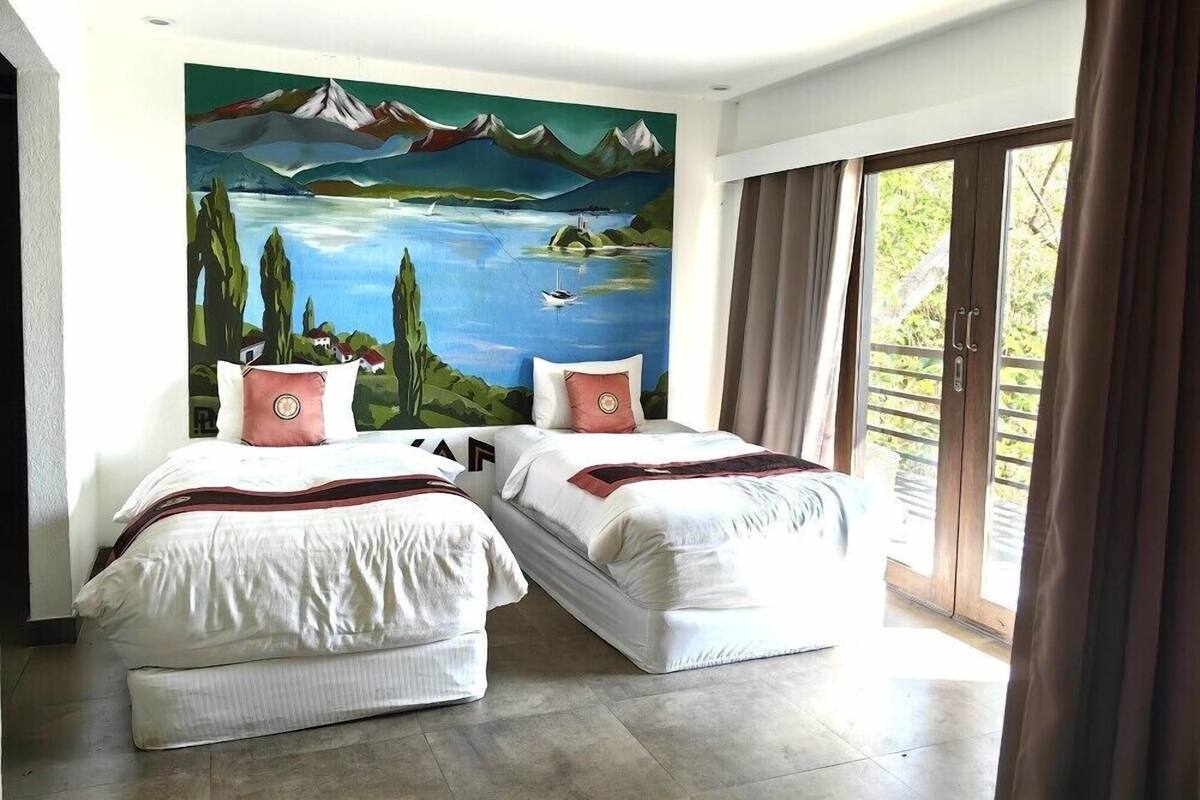 Une chambre avec deux lits, une fresque représentant un paysage et une grande fenêtre avec des rideaux.