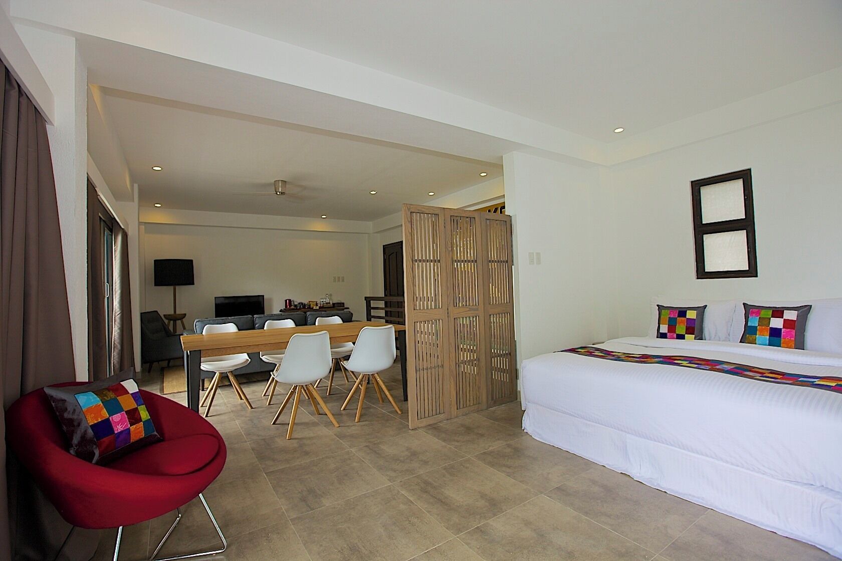 A modern hotel room with a large bed, a dining area with a wooden table and chairs, and a red chair.