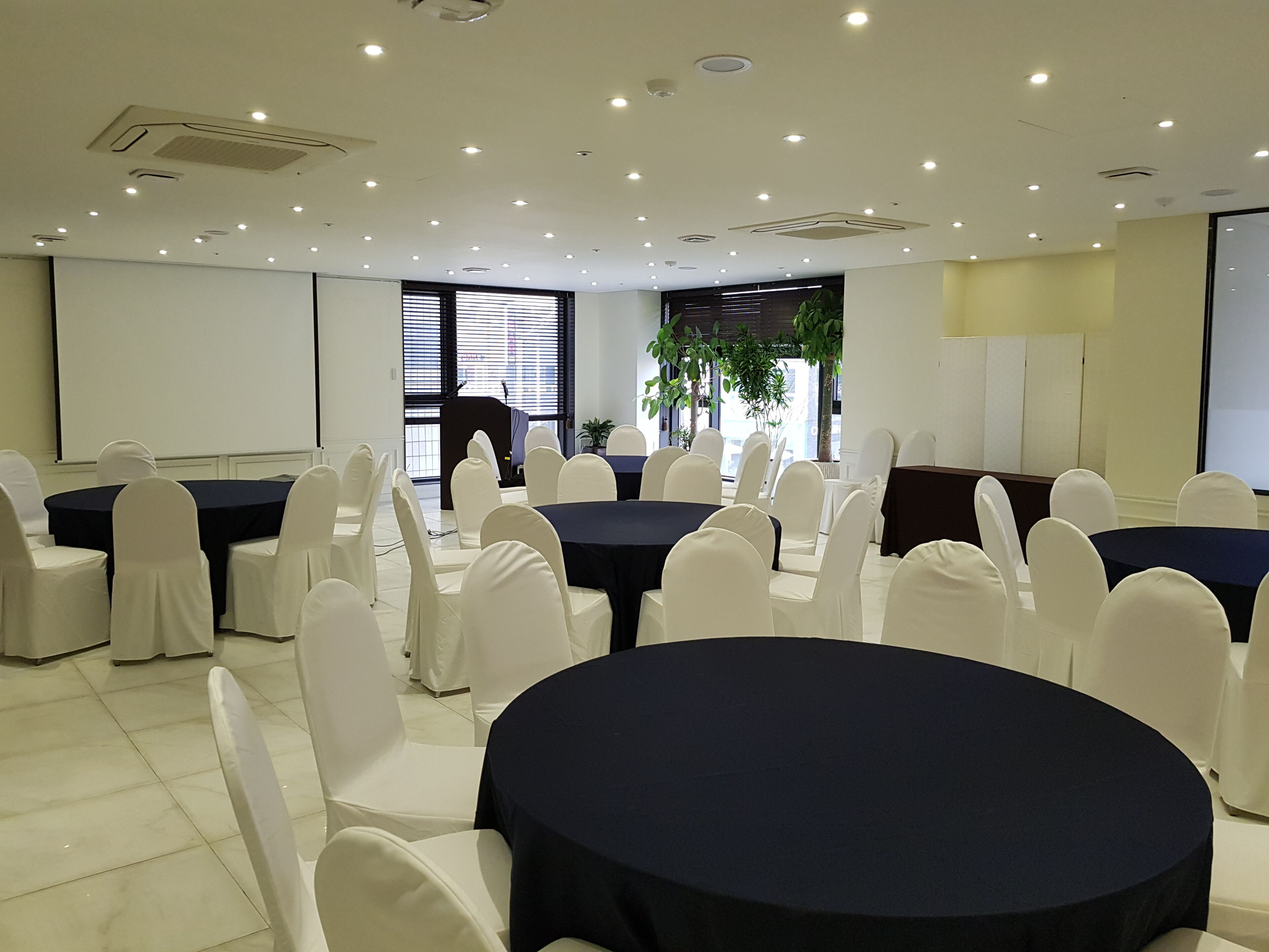 Un salón de conferencias con mesas redondas, sillas blancas y una pantalla para proyector.