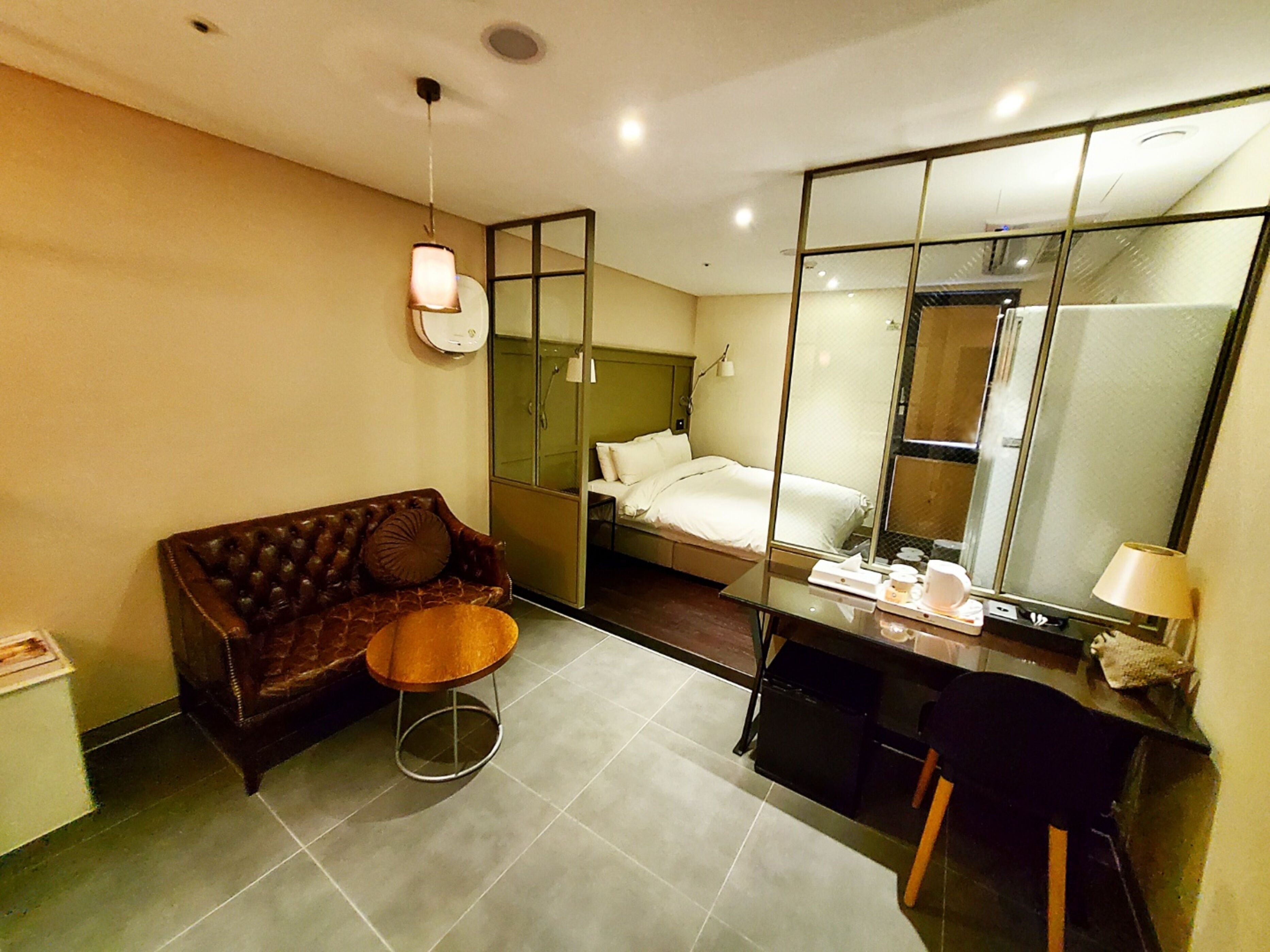 Habitación de hotel compacta con un sofá de cuero marrón, una mesita redonda pequeña, un escritorio con una silla, un espejo grande y una cama con ropa blanca.