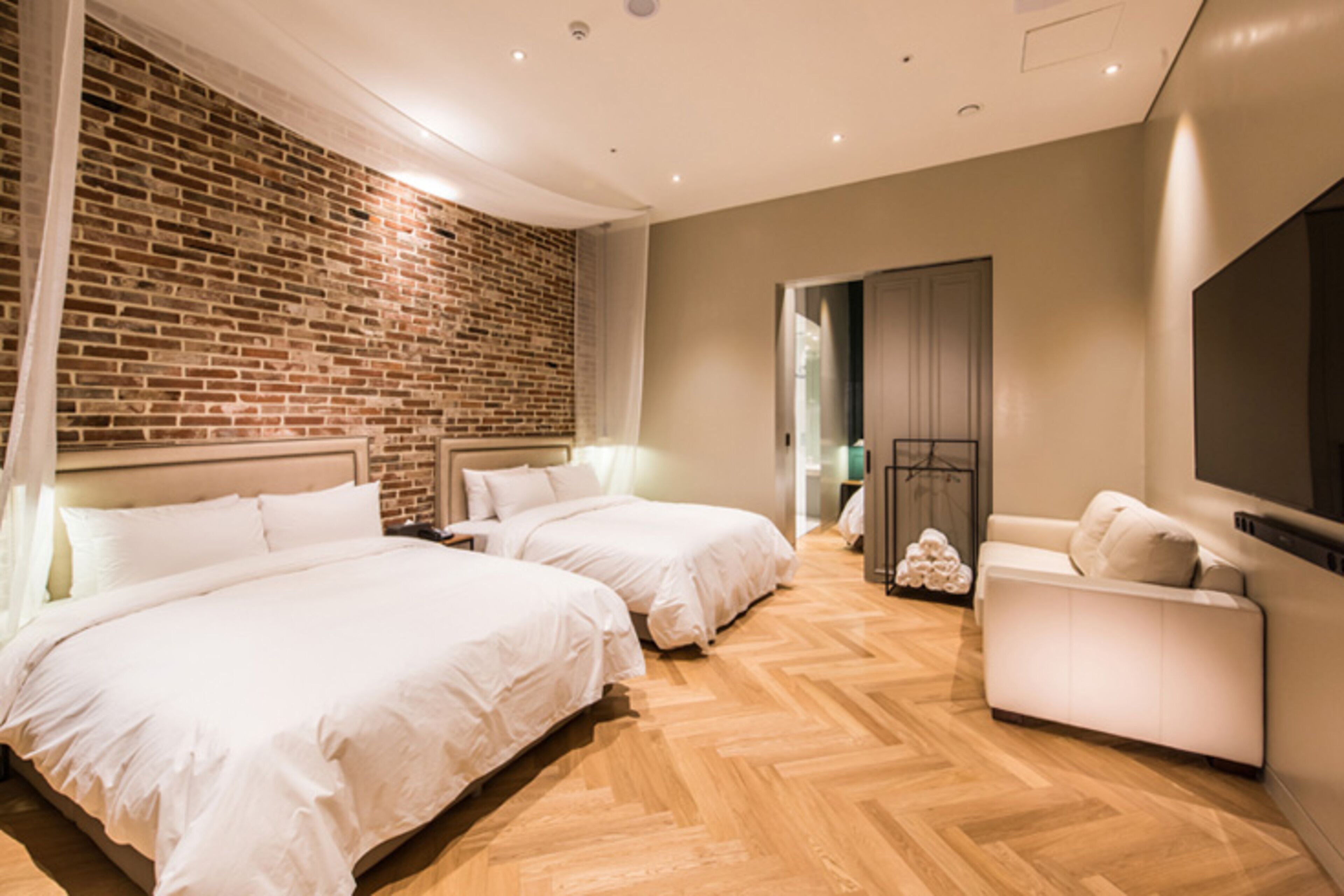 A modern hotel room with two beds, a sofa, a flat-screen TV, and a brick accent wall.