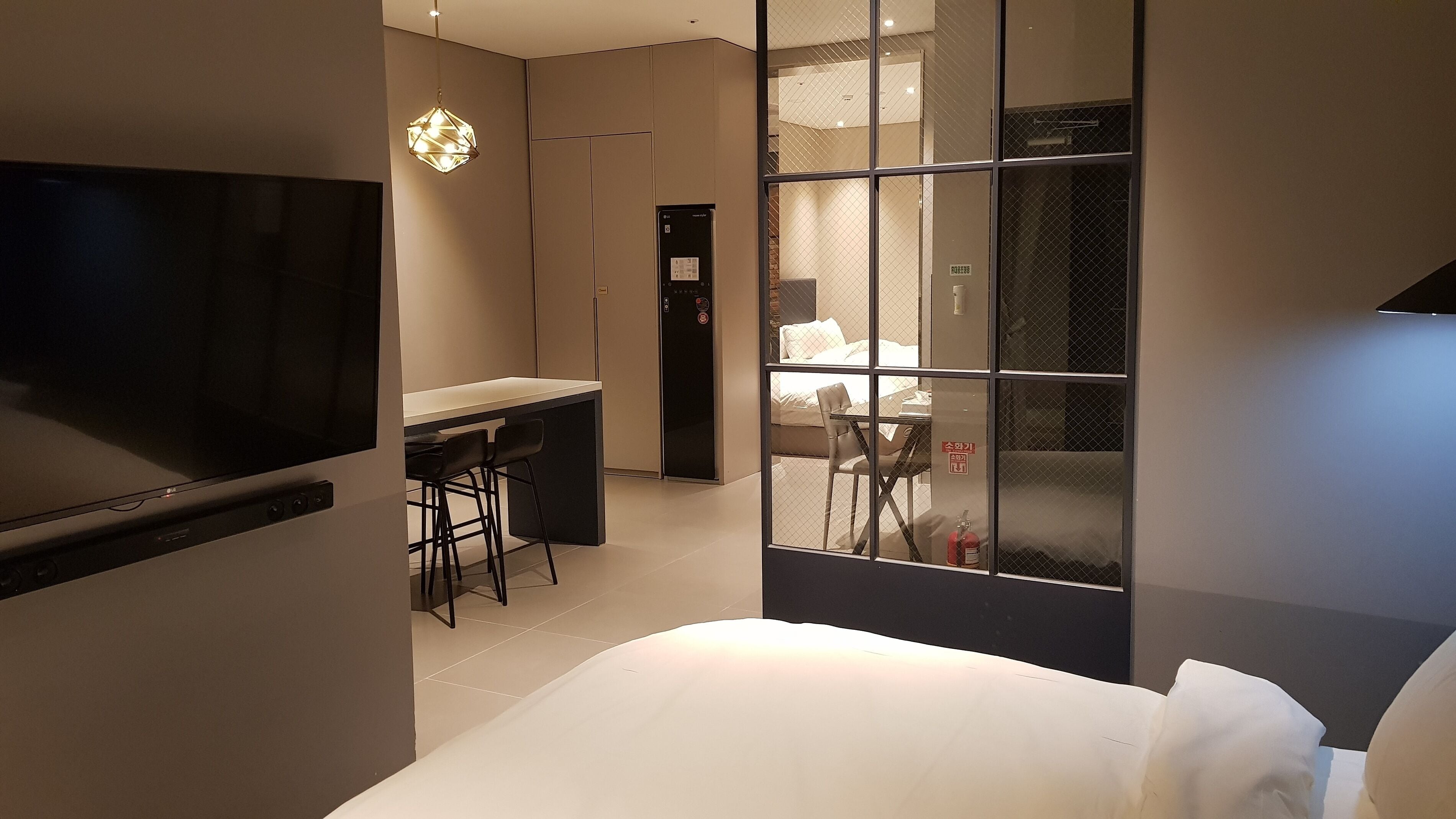 A modern hotel room with a bed, desk, and chairs. There is a TV on the left, a window with a view of the outside, and a door leading to another room.