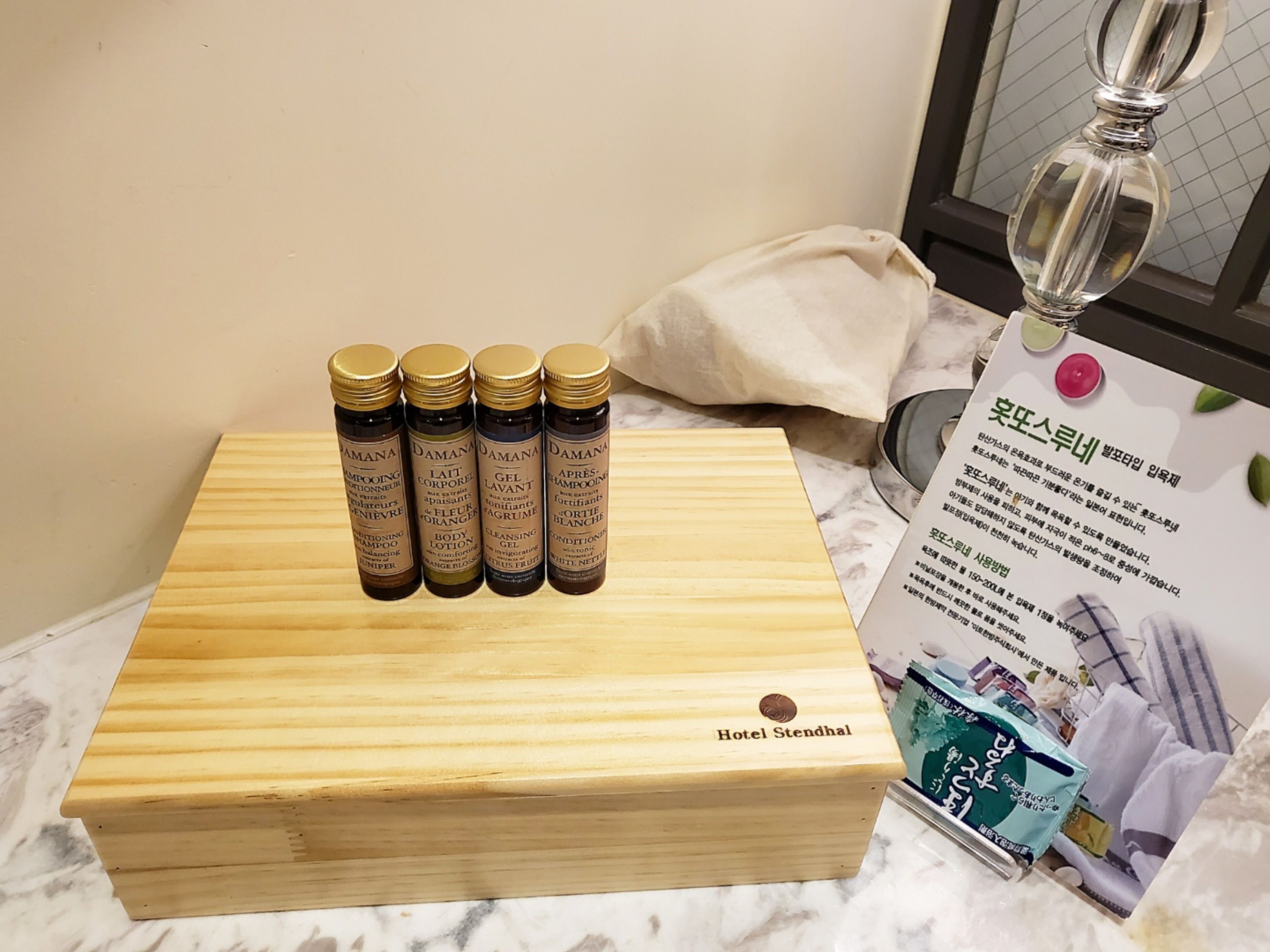 Una caja de madera con la etiqueta 'Hotel Stendhal', cuatro botellas de productos para el cabello y un folleto.
