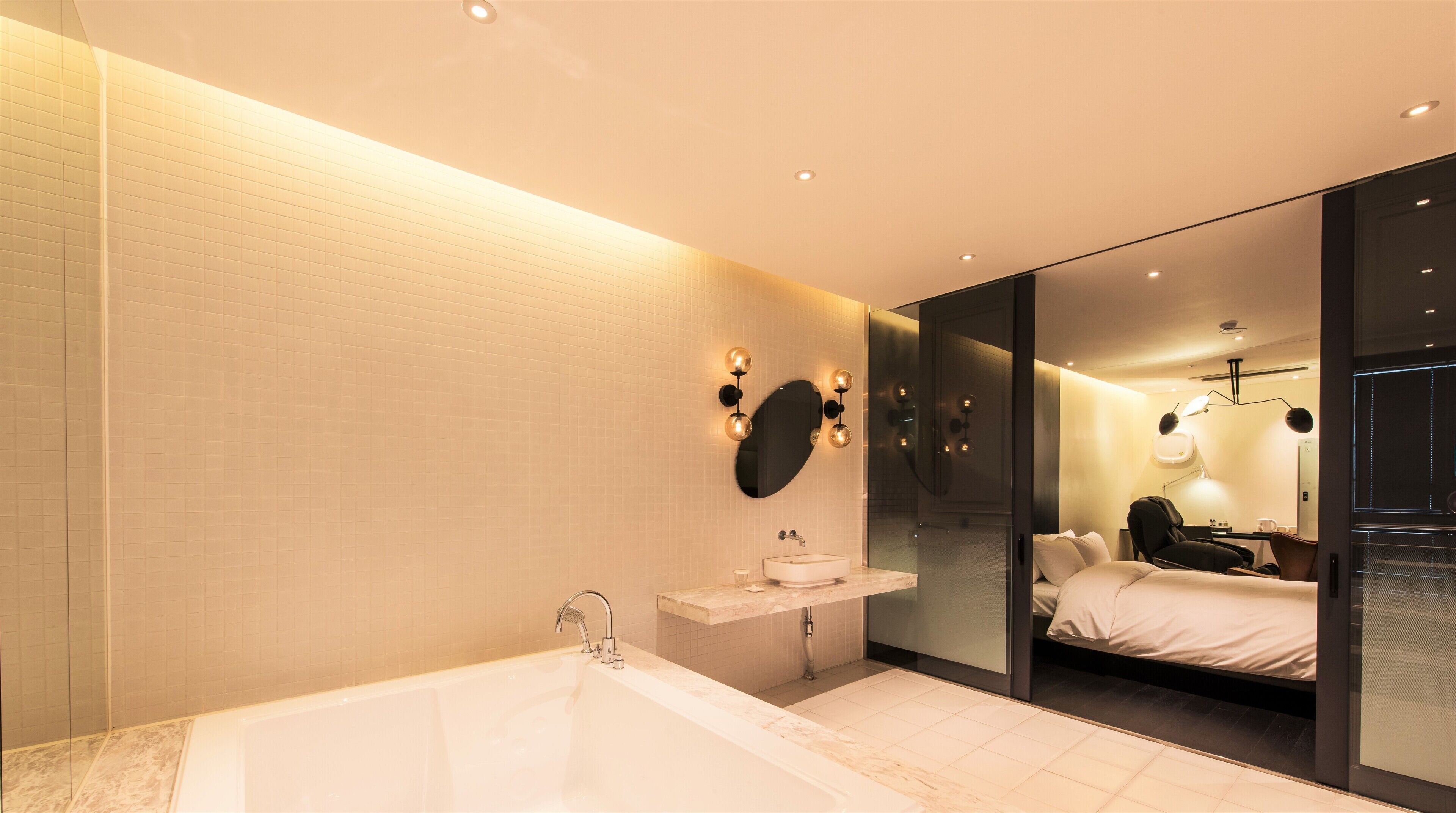 Une salle de bain moderne avec une grande baignoire, un meuble-lavabo et un miroir. Une cloison en verre sépare la salle de bain d’une chambre à coucher qui contient un lit et une table de chevet.