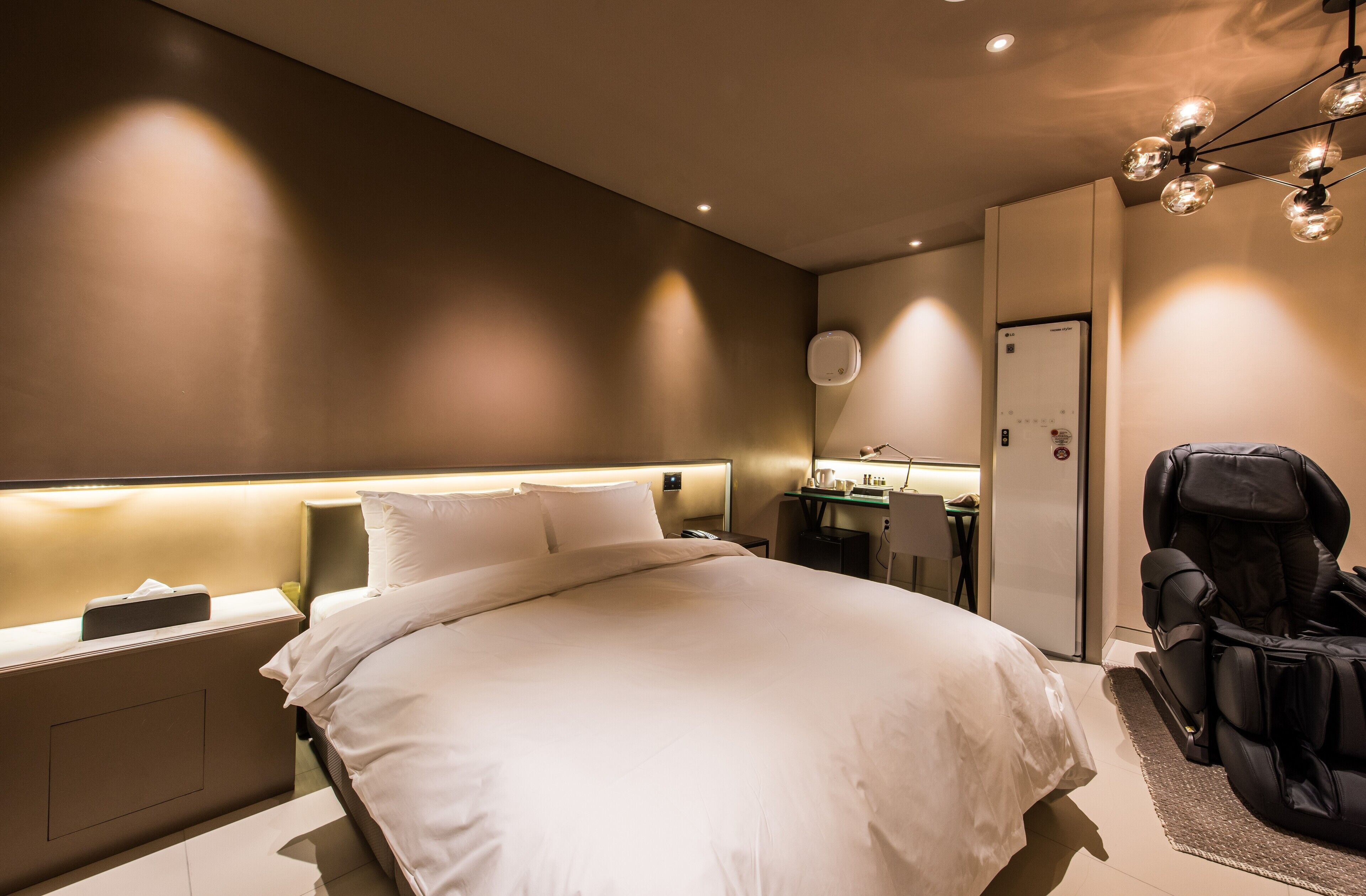 Una habitación de hotel moderna con una cama grande, mesitas de noche, un escritorio con silla, un sillón de masaje y un minibar.