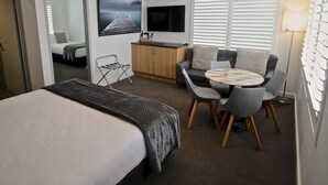 A hotel room with a bed, a desk, a chair, a sofa, a TV, and a window with blinds. - Quays Hotel (Batemans Bay)