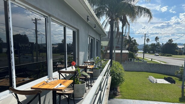 A hotel patio with outdoor seating, large windows, and a view of the surrounding area including a road and palm trees. - Quays Hotel (Batemans Bay)