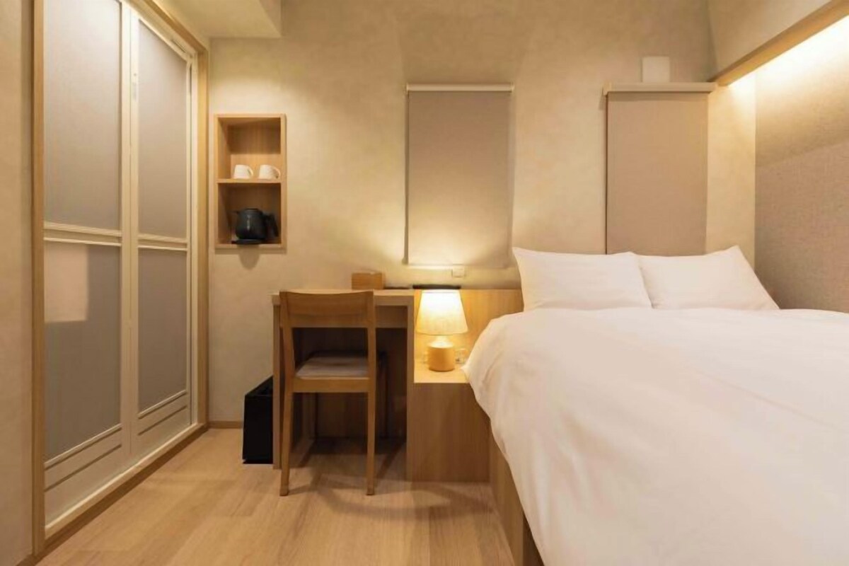 A hotel room with a bed, a desk with a chair, a nightstand with a lamp, and a shelf with cups and a pot.