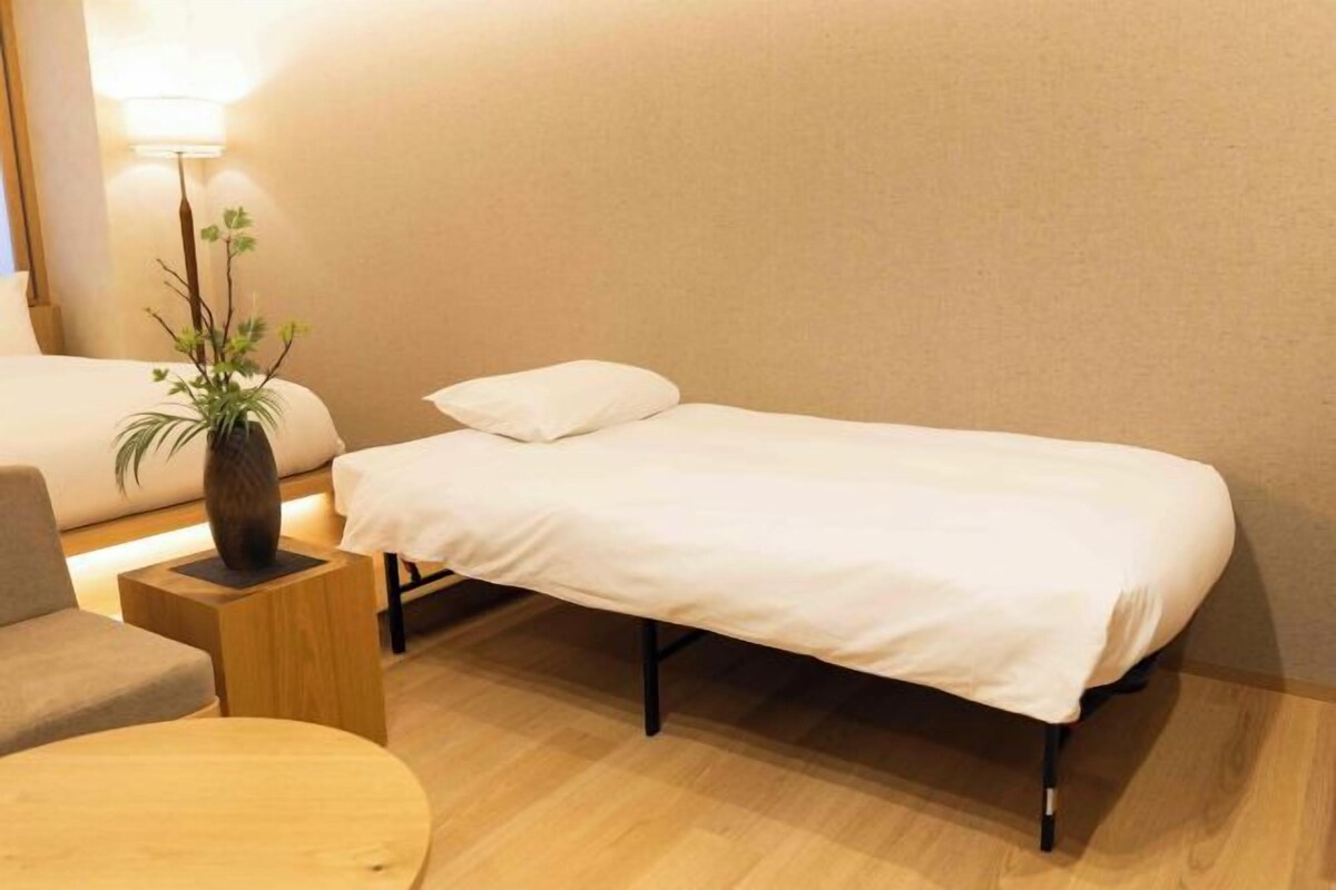 A single bed with a white mattress and pillows, a small wooden nightstand with a vase, a round wooden table, and a lamp.