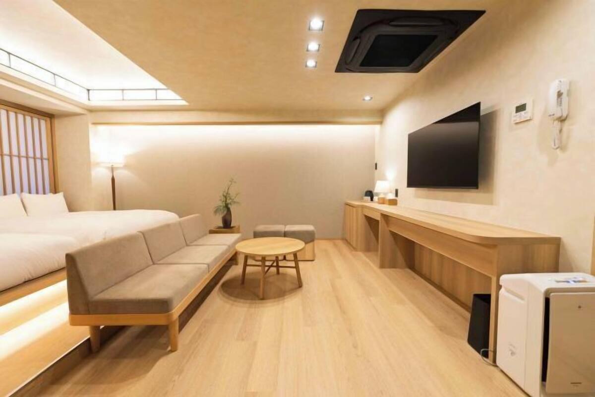A compact hotel room with a sofa, a small round table, a TV mounted on the wall, and a desk with a computer.