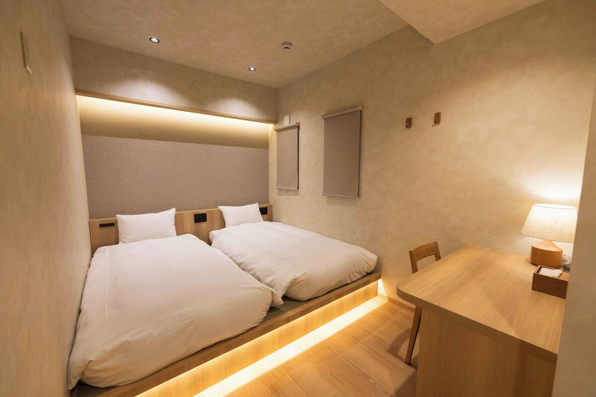 A hotel room with two beds, a wooden desk, and a small table with a lamp.