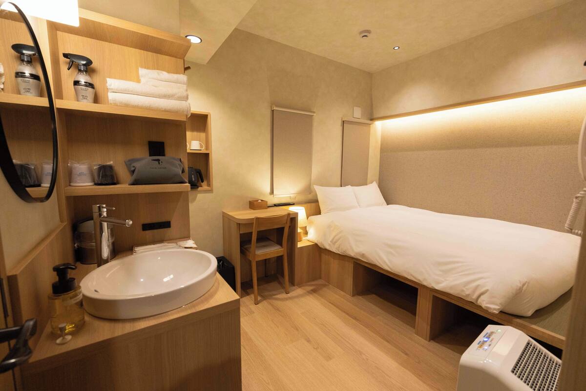 A hotel room with a large bed, a bedside table with a lamp, a wooden nightstand, a large round sink, and a mirror.