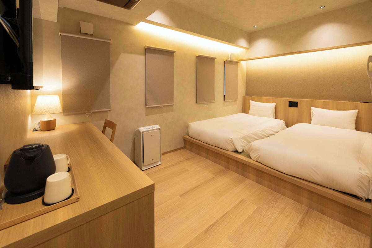 A hotel room with a wooden desk, two beds, a bedside table, a lamp, and a heater.