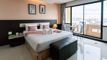 A hotel room with a large bed, a bedside table, a nightstand, a window with a city view, and a small balcony with a table and chairs.