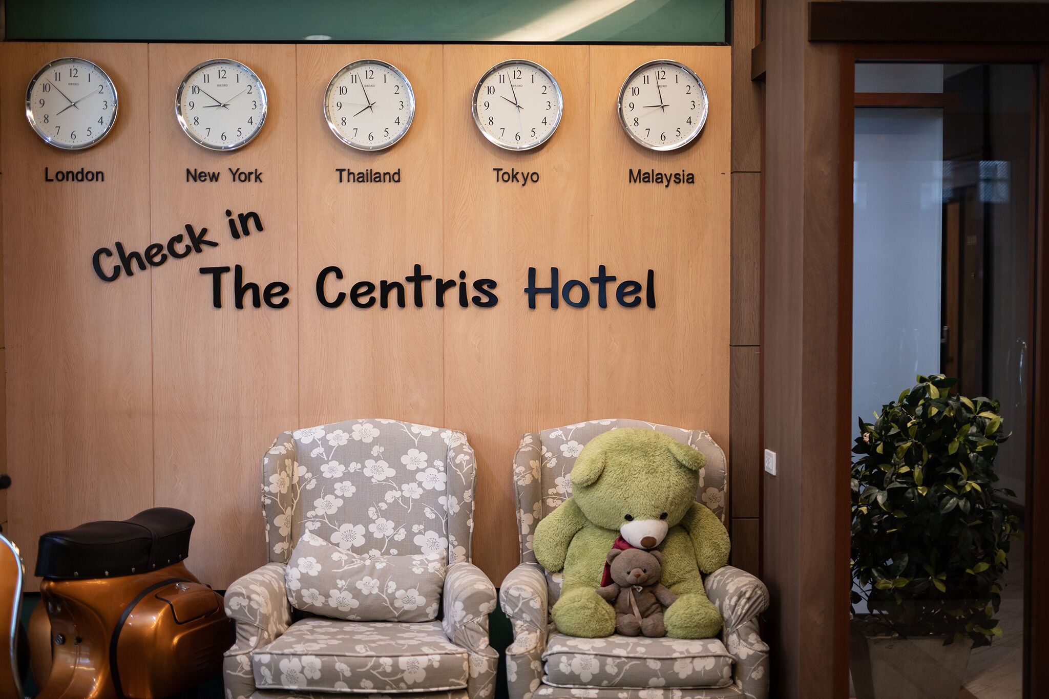 Zona de recepción con relojes mostrando horarios para Londres, Nueva York, Tailandia, Tokio y Malasia. Hay dos sillones florales y un osito de peluche grande de color verde.