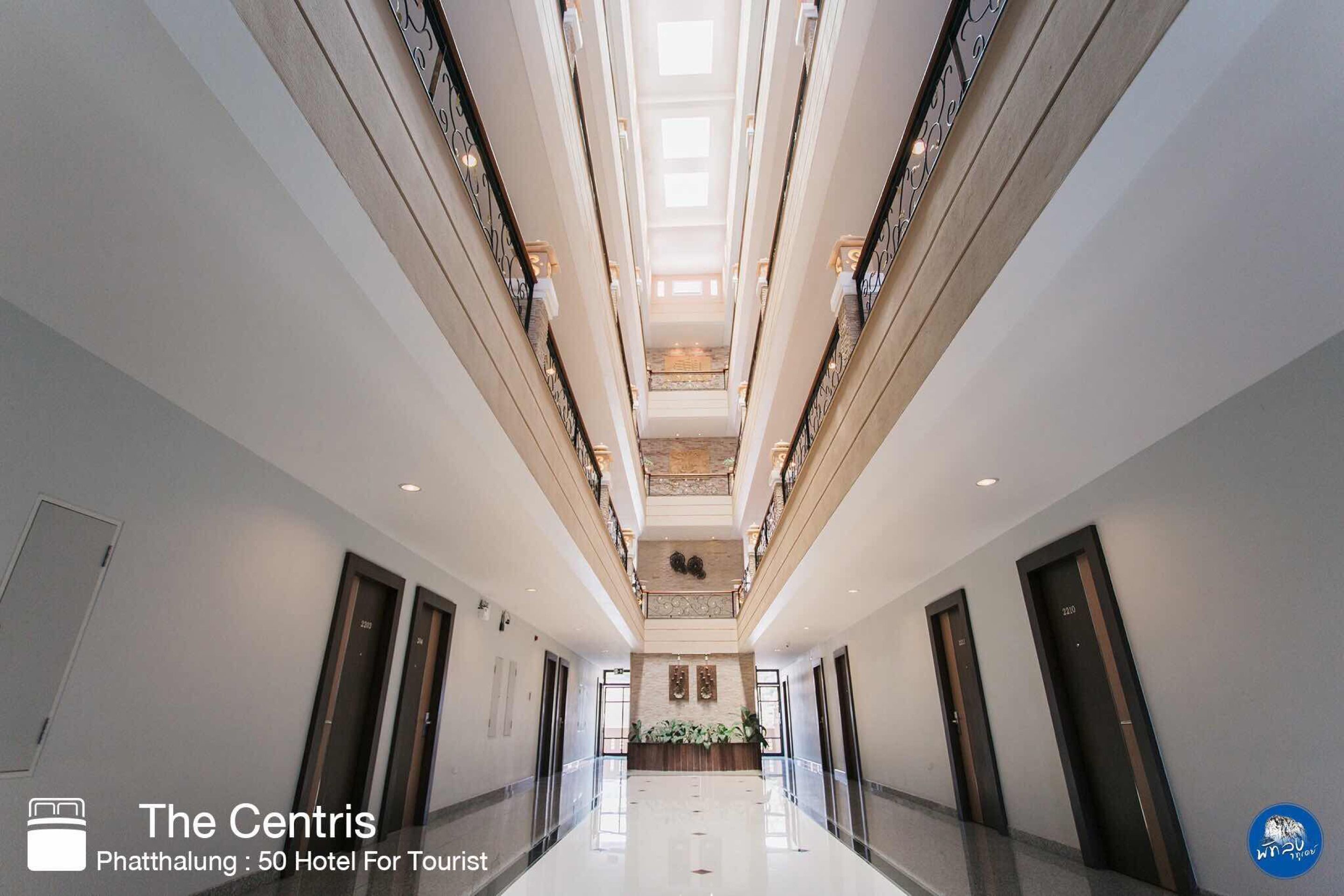 A modern hotel corridor with a central staircase, multiple doors, and a reception area at the end.